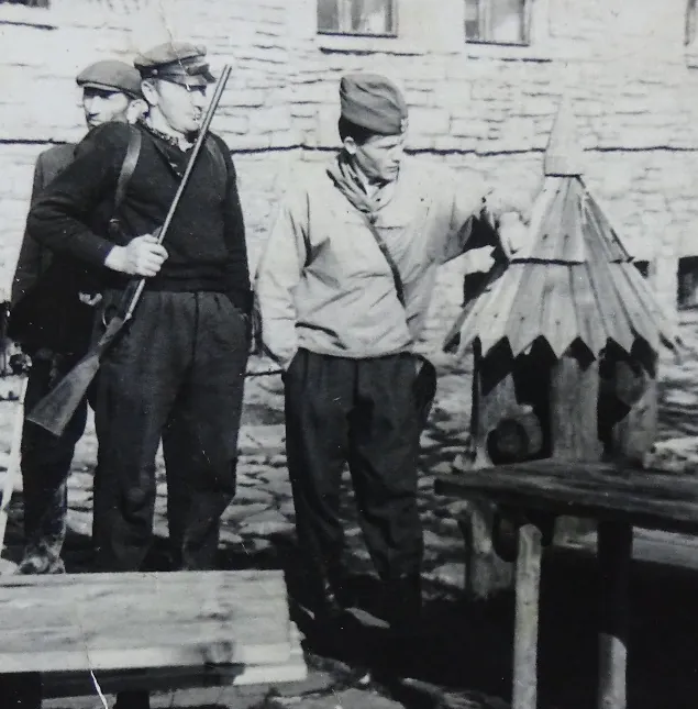 Archiwalna czarno-biała fotografia: trzej mężczyźni w kurtkach i furażerkach przed kamiennym budynkiem, jeden trzyma karabin