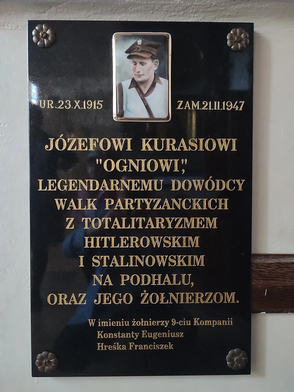 Czarna tablica pamiątkowa ze zdjęciem Józefa Kurasia i złotym napisem „Józefowi Kurasiowi »Ogniowi«, legendarnemu dowódcy walk partyzanckich z totalitaryzmem hitlerowskim i stalinowskim na Podhalu, oraz jego żołnierzom”